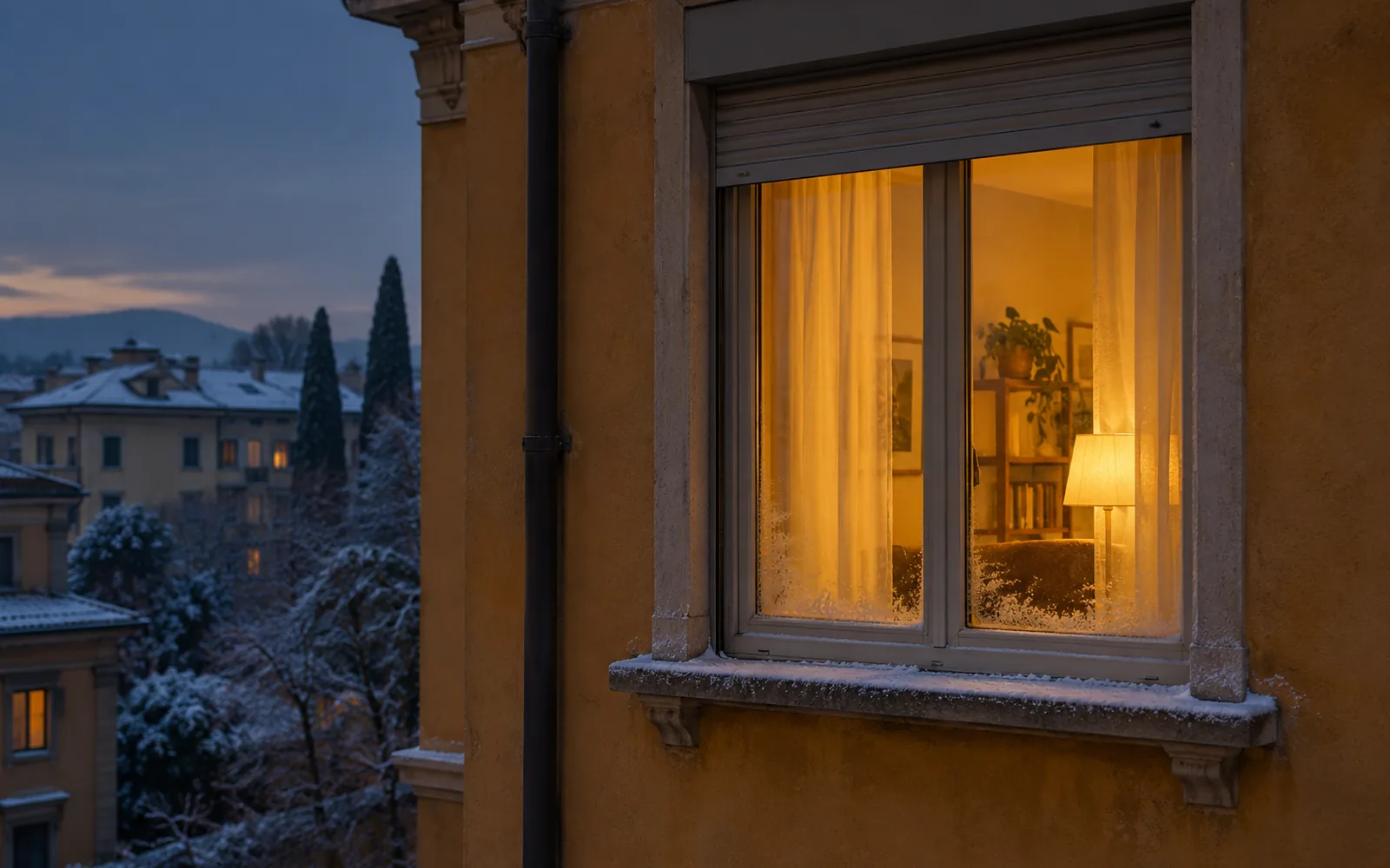 Finestra di un palazzo color ocra vista da fuori in una sera d’inverno, con la luce calda dell’interno, il cassonetto della tapparella sopra e la brina sul davanzale