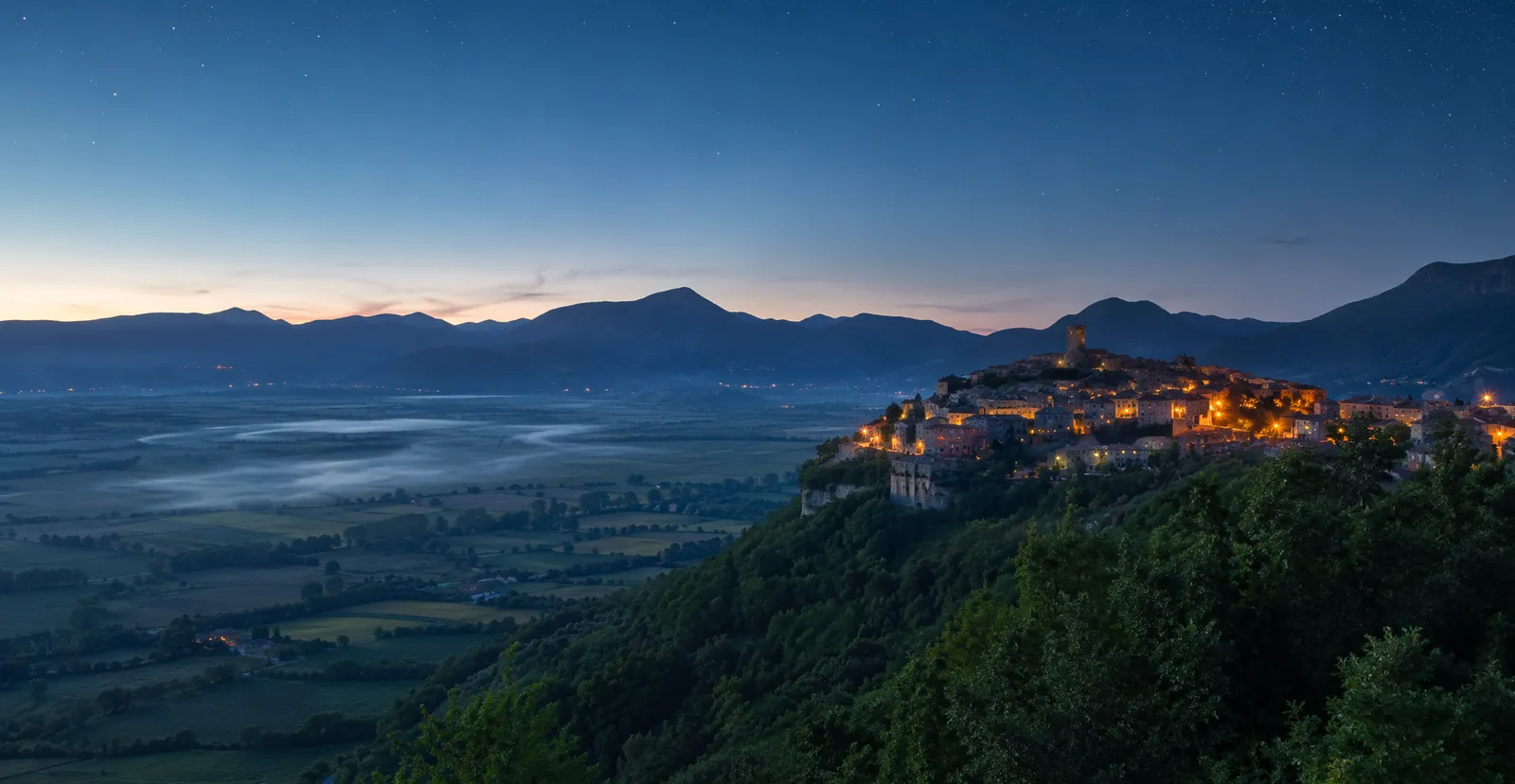 Borgo ciociaro in collina al crepuscolo, con la valle sottostante coperta da banchi di nebbia serale