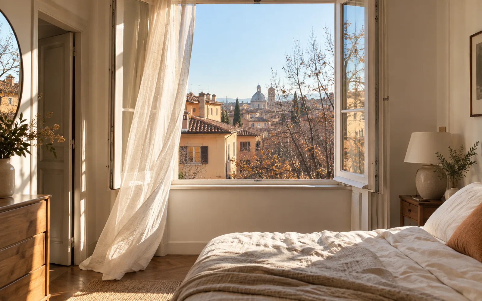 Finestra di una camera aperta del tutto verso l’interno in una mattina d’autunno, con la tenda di lino mossa dall’aria e i tetti della città fuori
